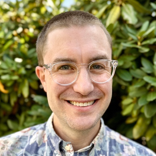 photograph focused on a smiling man with pale skin, buzzed short
          hair, blue eyes, and glasses wearing a light-colored patterned shirt
          and standing in front of green bush